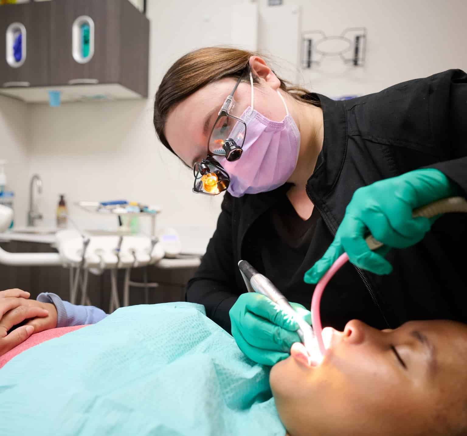 Harrison Family Dental team member wearing lighted loupes cleaning a patient’s teeth while using suction