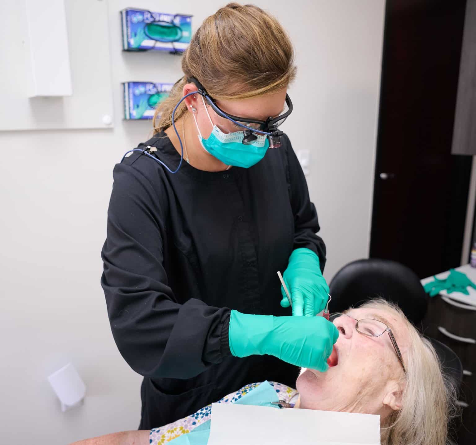 Harrison Family Dental team member wearing loupes examining and cleaning an older patient’s teeth in the dental chair
